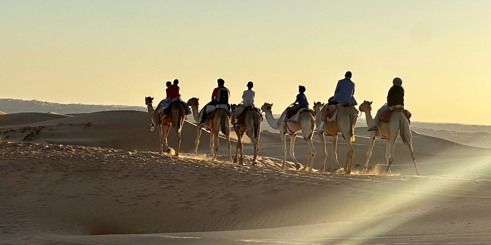 Camel caravan crossing desert dunes at sunset, riders following a single track toward the horizon