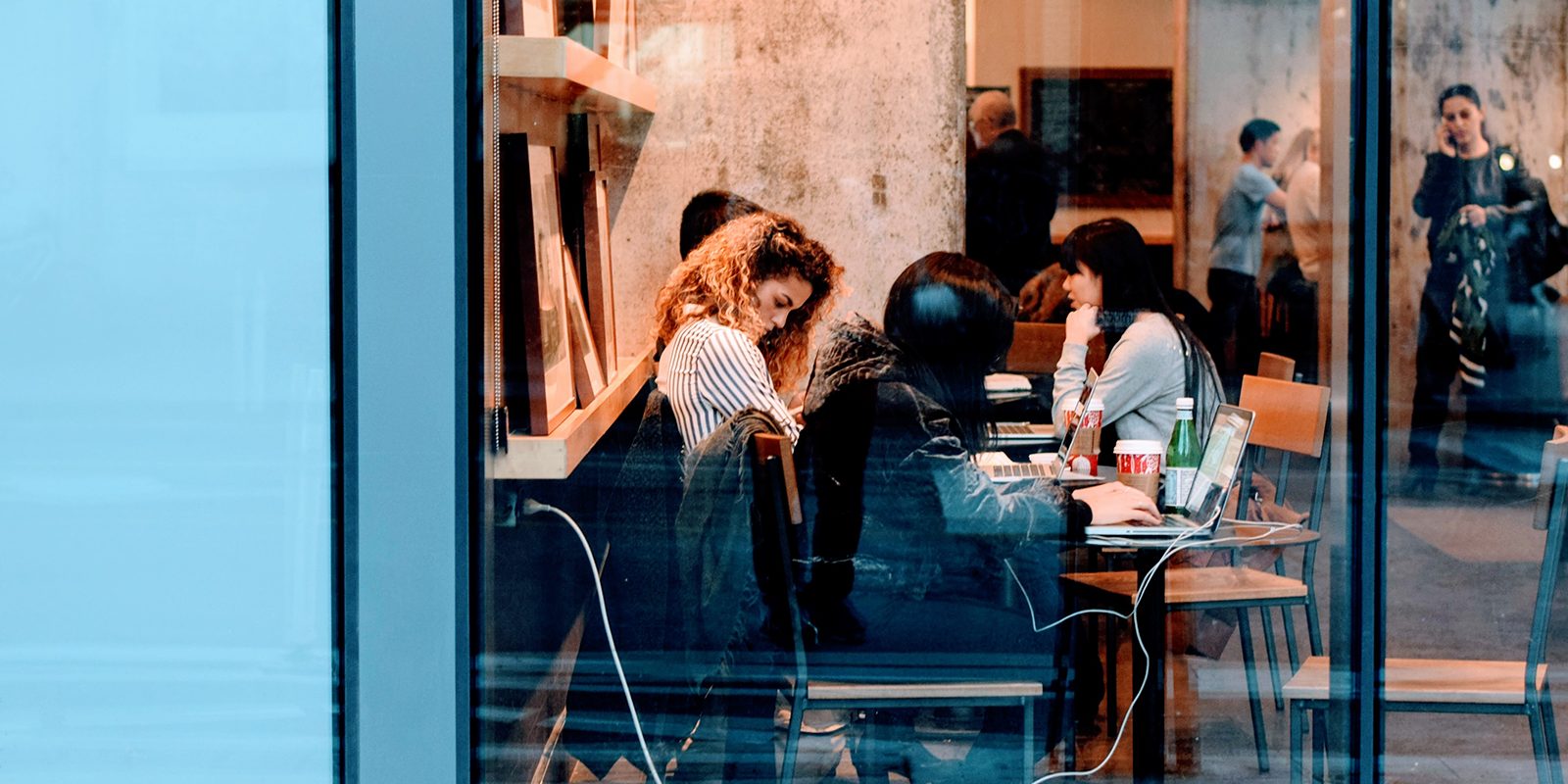 People talking over laptops at a cafe table, seen through a window